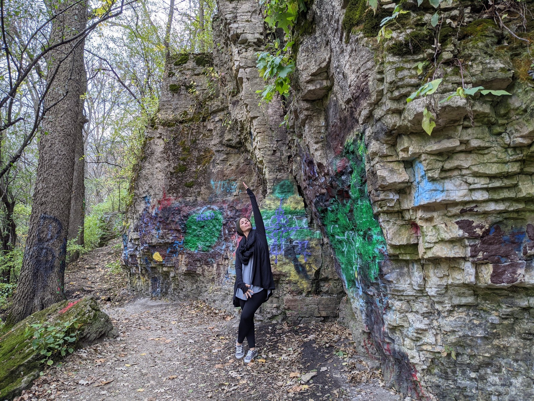 Liz stretching one arm skyward on a leaf-covered trail beside a painted limestone bluff